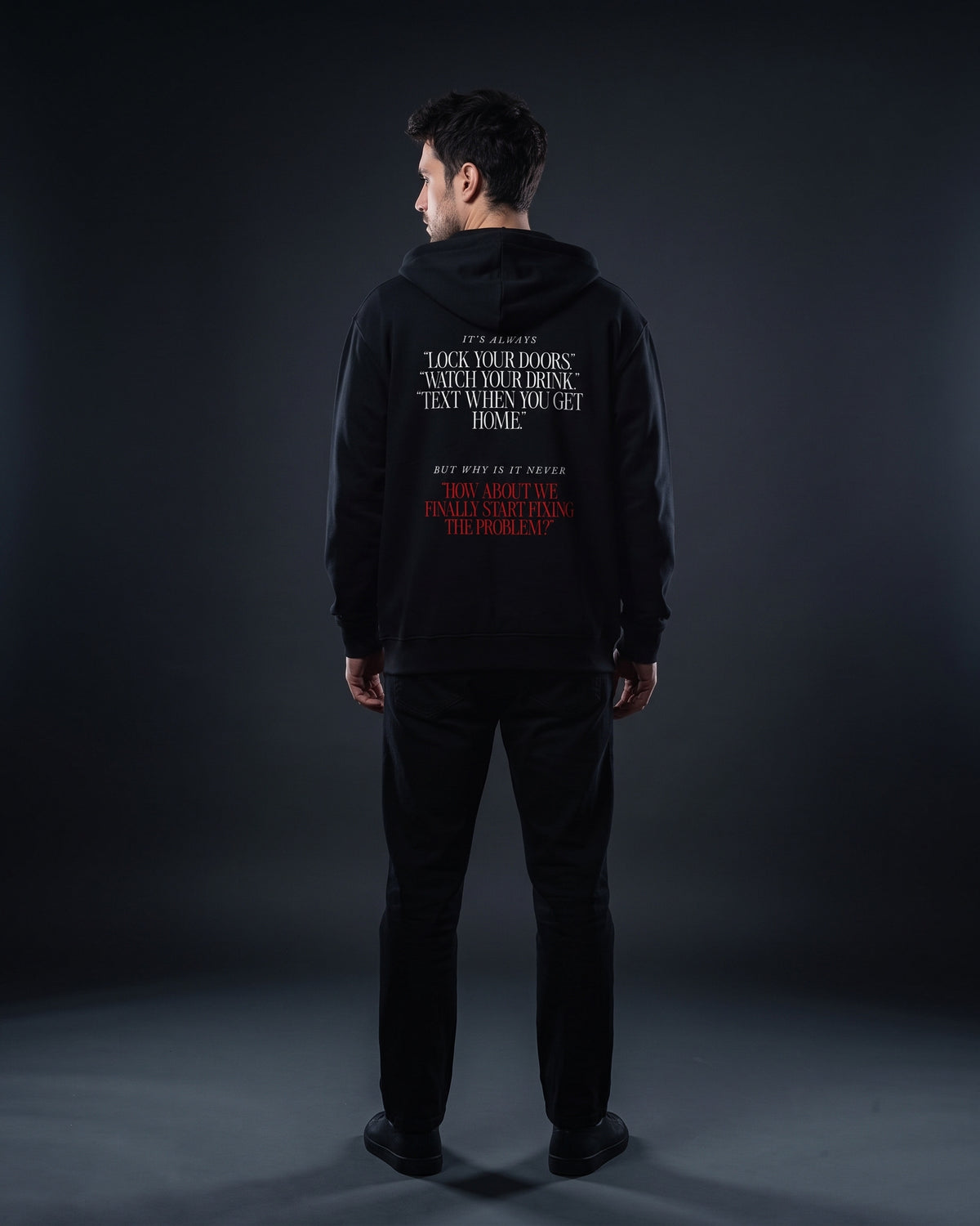 Mann in schwarzem Zip-Hoodie von hinten, Rückenprint „It's always ‚Lock your doors.‘ ‚Watch your drink.‘ ‚Text when you get home.‘ But why is it never ‚How about we finally start fixing the problem?‘“