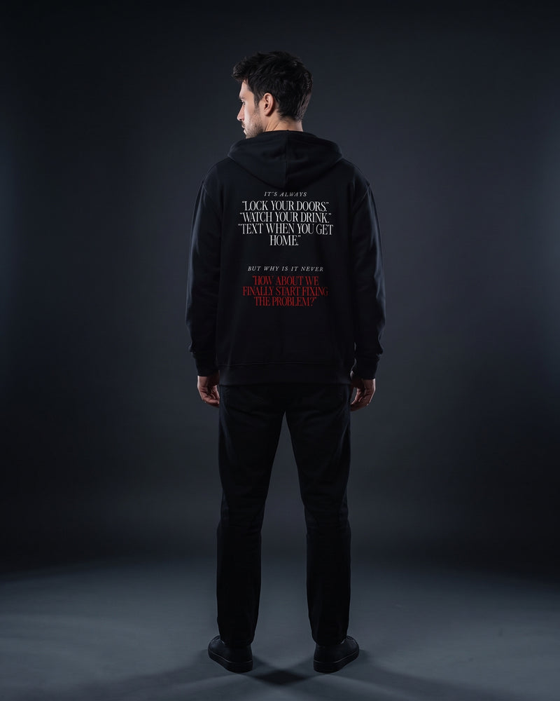 Mann in schwarzem Zip-Hoodie von hinten, Rückenprint „It's always ‚Lock your doors.‘ ‚Watch your drink.‘ ‚Text when you get home.‘ But why is it never ‚How about we finally start fixing the problem?‘“